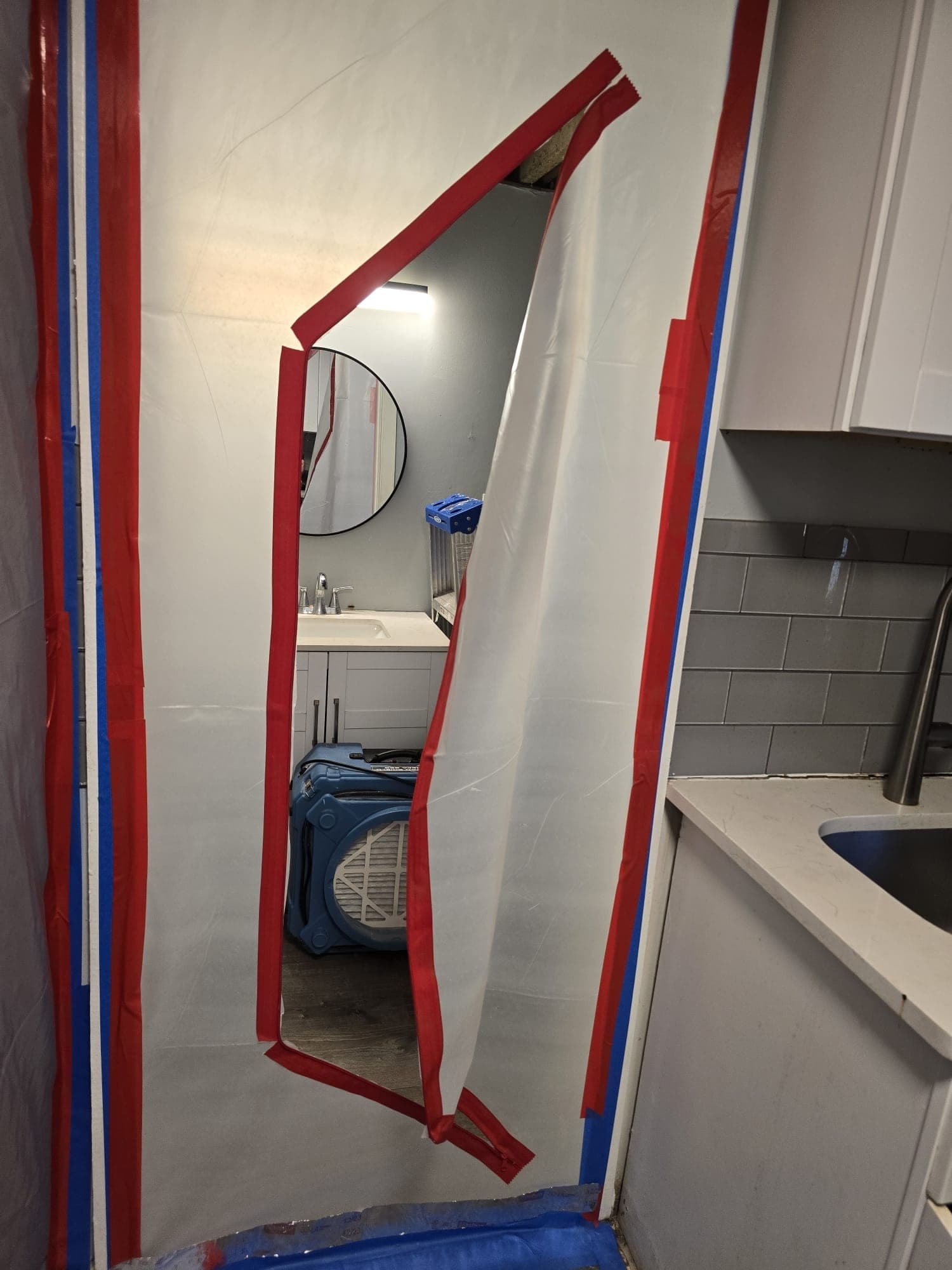 Poly sheeting containment doorway with red-taped zipper opening into a bathroom where an air scrubber runs
